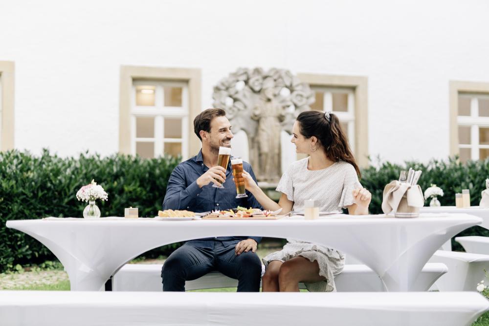 Mann und Frau am Tisch im Garten stoßen mit Bier an